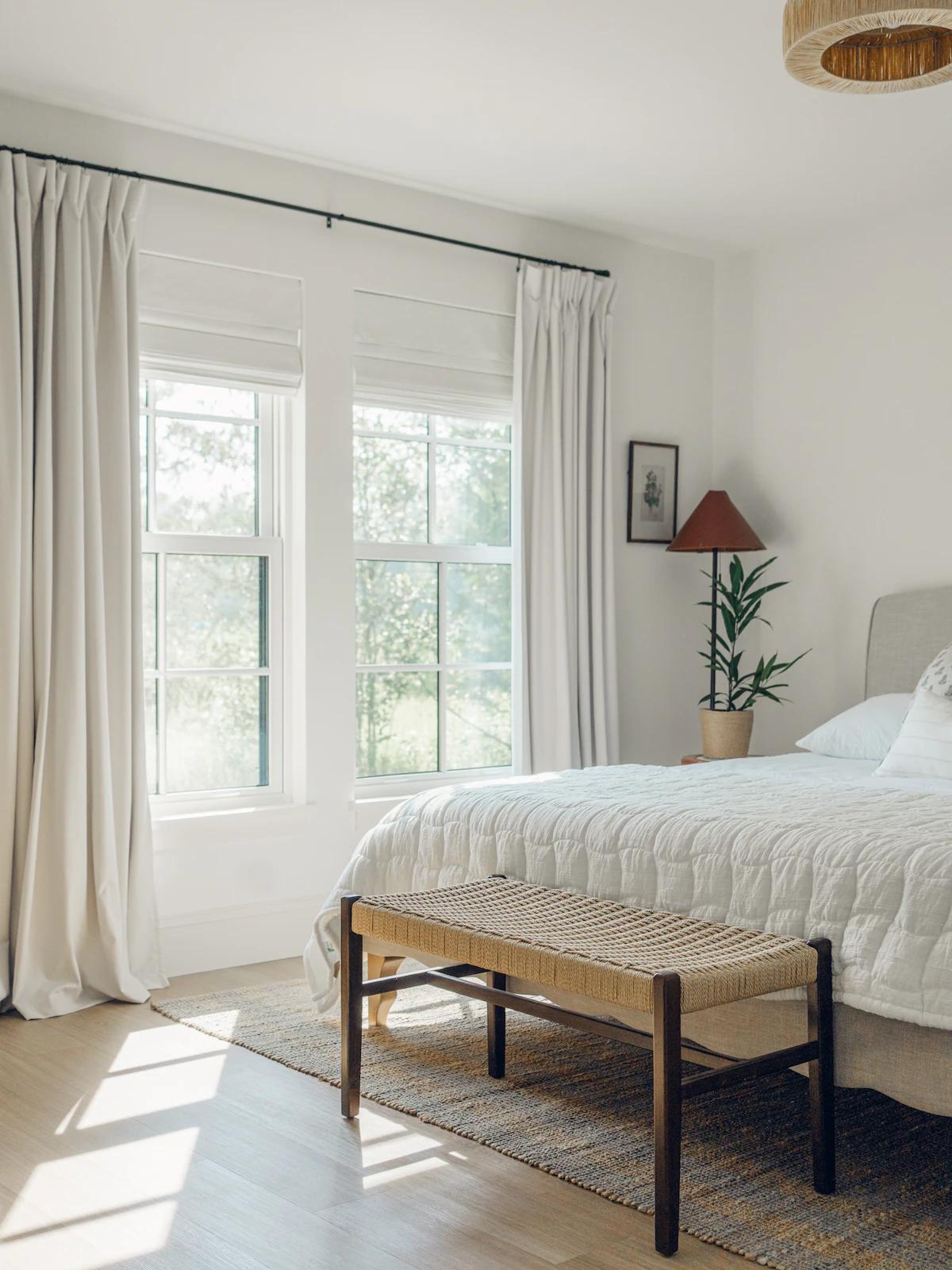 Sunlit vacation rental bedroom with white bedding, a woven bench at the foot of the bed and tall windows